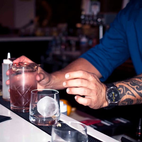 A bartender with tattooed arms prepares a cocktail with a large ice sphere at Byblos.