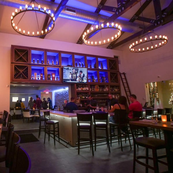 Byblos restaurant's bar area with patrons, a TV, and illuminated liquor shelves under large chandeliers.