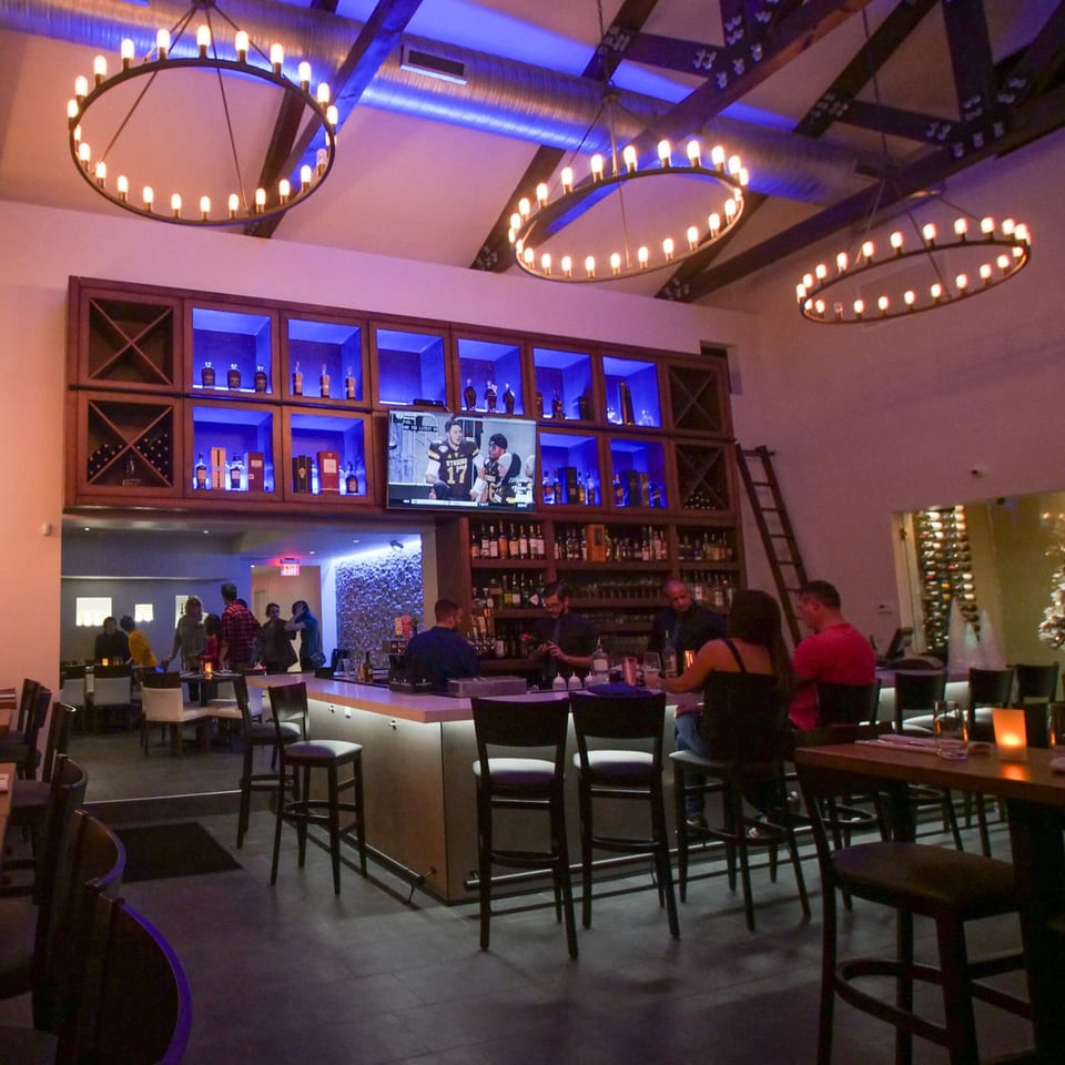 Byblos restaurant's bar area with patrons, a TV, and illuminated liquor shelves under large chandeliers.