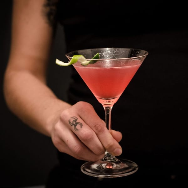 A hand with a spider tattoo holds a martini glass filled with a pink cocktail and lime garnish.