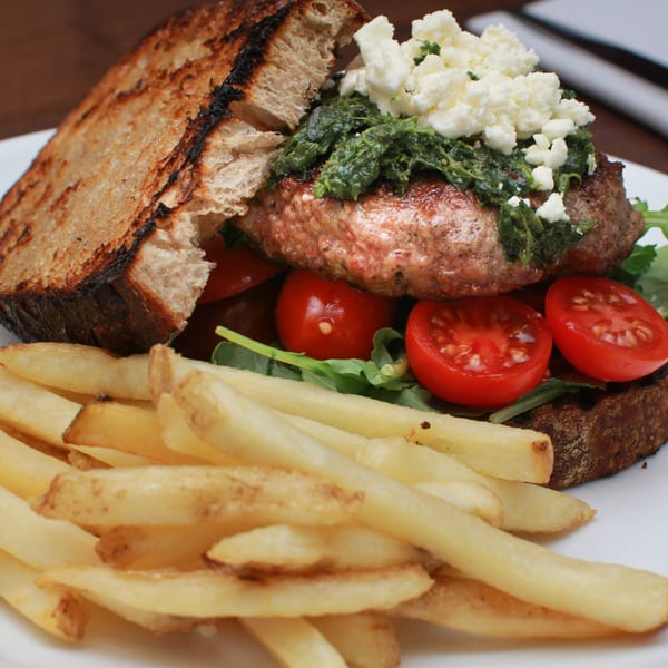 Byblos Mediterranean burger with pesto, feta, tomatoes, arugula, and fries on a white plate.