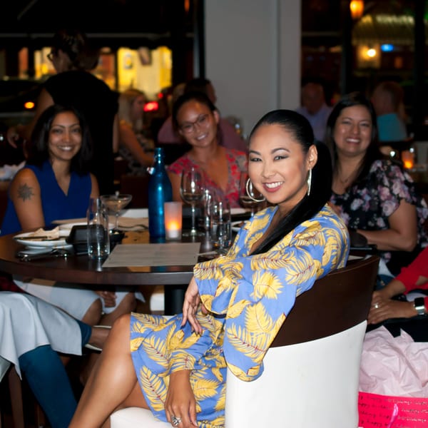 Smiling woman in a blue and yellow dress seated at a table with friends at Byblos.