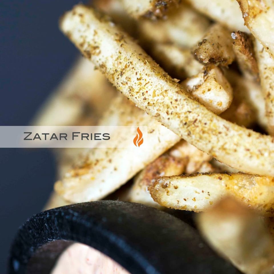 Close-up of golden-brown Zatar fries, seasoned with spices, in a dark container.